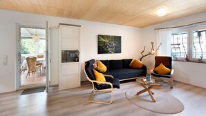 A modern living room with a dark sofa, two armchairs, a glass coffee table, and a wooden ceiling.