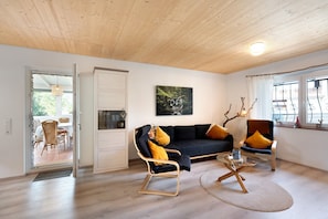 A modern living room with a dark sofa, two armchairs, a glass coffee table, and a wooden ceiling.