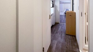 A narrow hallway with white walls and wooden flooring, leading to a kitchen area with a microwave and refrigerator.