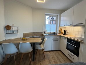 A compact kitchen with a wooden dining table, two chairs, a built-in oven, and a window with blinds.