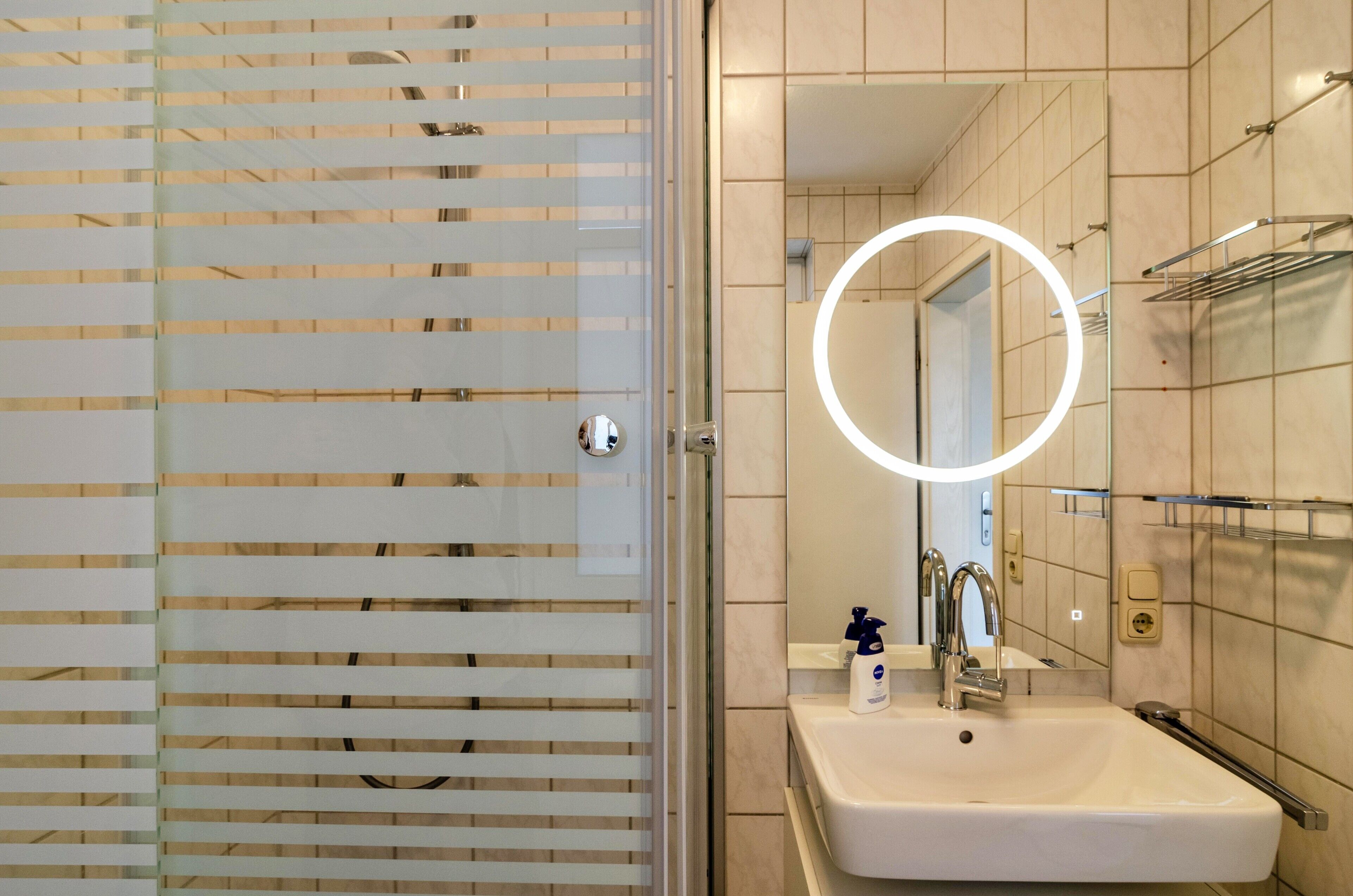 A modern bathroom with a glass door, a sink, a mirror with a circular light, and a towel rack.
