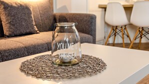 Ein klares Glasgefäß mit der Aufschrift 'YOU ME & THE SEA' auf einem weißen Tisch, umgeben von einer gewebten Tischmatte, in einem Raum mit grauem Sofa und weißen Stühlen.
