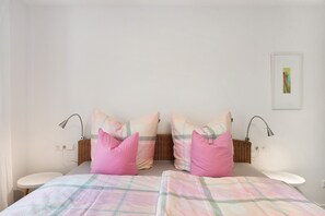 A bedroom with two beds, a bedside table, a wall-mounted lamp, a wicker basket, and a framed picture on the wall.