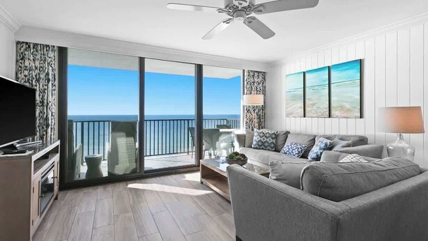 Une chambre d’hôtel moderne avec un balcon, un canapé gris, un téléviseur à écran plat et une grande fenêtre donnant sur la mer.