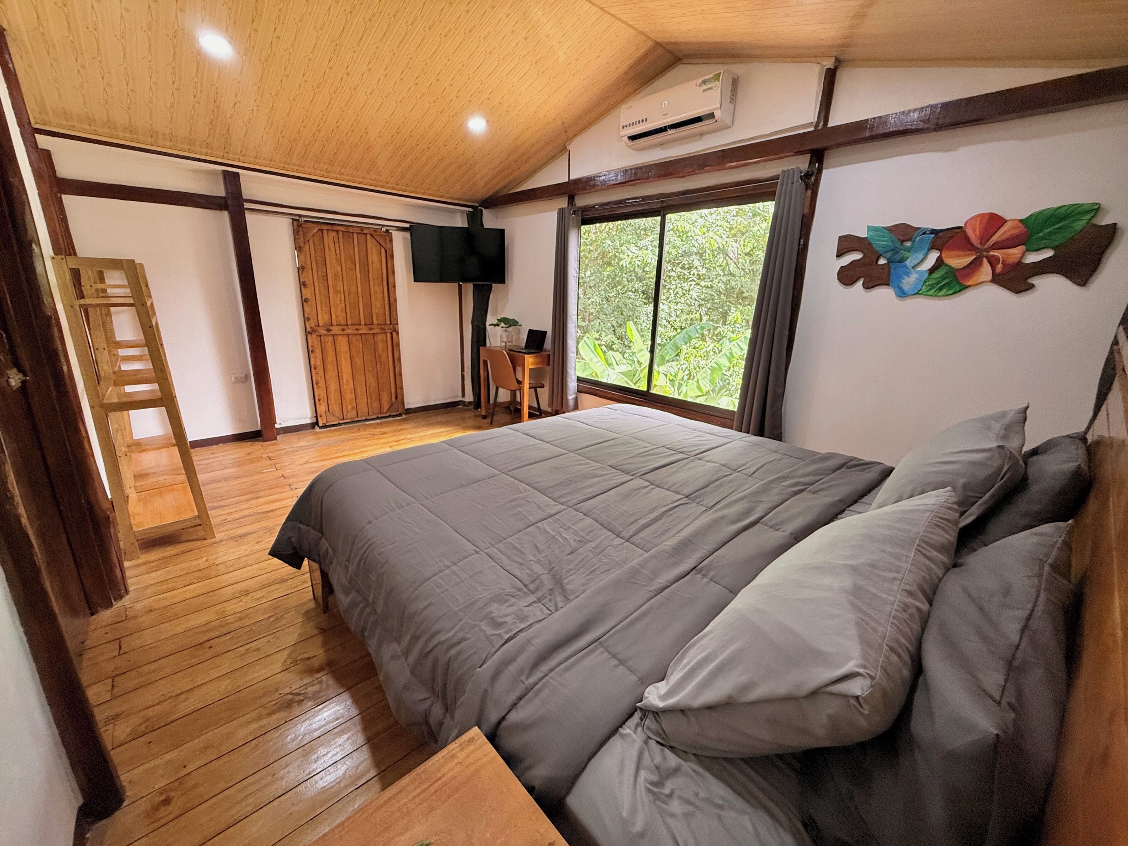 A bedroom with a large bed, wooden flooring, a desk with a laptop, a TV, and a window with a view of greenery.
