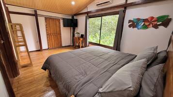 A bedroom with a large bed, wooden flooring, a desk with a laptop, a TV, and a window with a view of greenery.