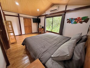 Une chambre avec un grand lit, un plancher en bois, un bureau avec un ordinateur portable, une télévision et une fenêtre donnant sur un paysage verdoyant.