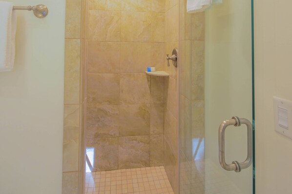 Une douche fermée par des parois en verre, avec des carreaux de couleur beige, une petite étagère et un porte-serviettes.