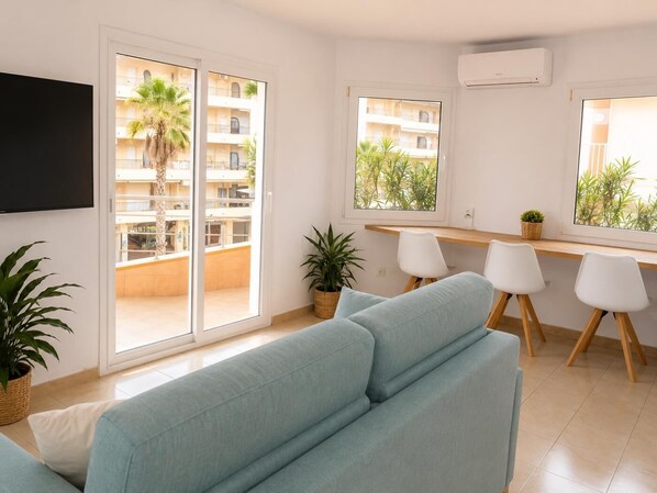 A modern living room with a sofa, a dining area with chairs, a TV, and a balcony with plants.