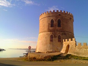 Ein historischer Turm mit rundbogigen Fenstern und einer befestigten Mauer in der Nähe eines Gewässers.