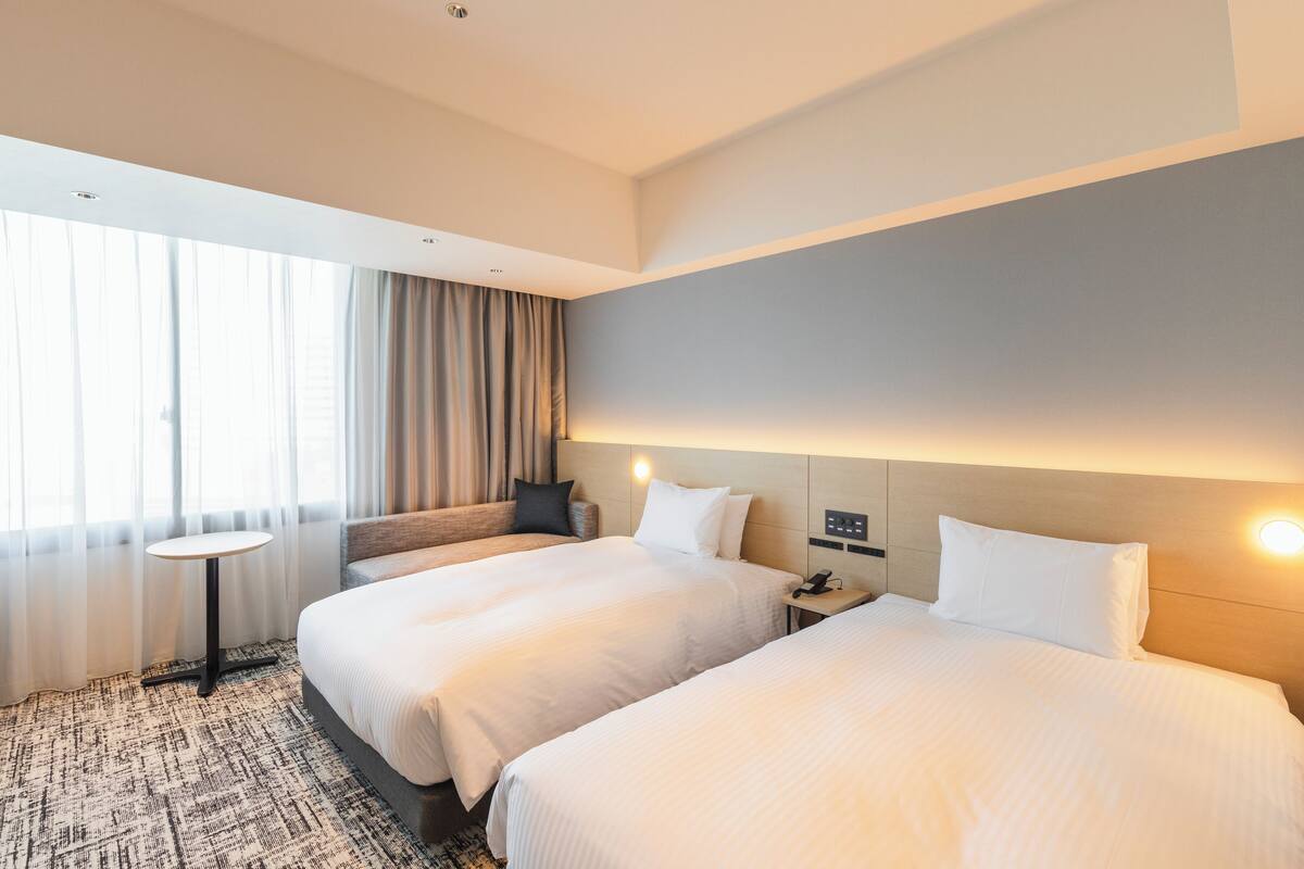 Habitación de hotel con dos camas, una mesita redonda pequeña, un sofá y un ventanal grande con cortinas.