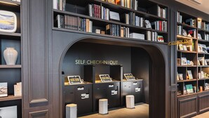 Ein Selbstcheck-in/Check-out-Bereich mit dunklen Holzelementen, mehreren Check-in-Terminals und einer Auswahl an Büchern sowie dekorativen Gegenständen auf Regalen.