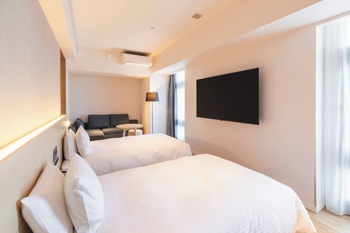 Habitación de hotel con dos camas, un televisor de pantalla plana montado en la pared, un escritorio y un sofá.