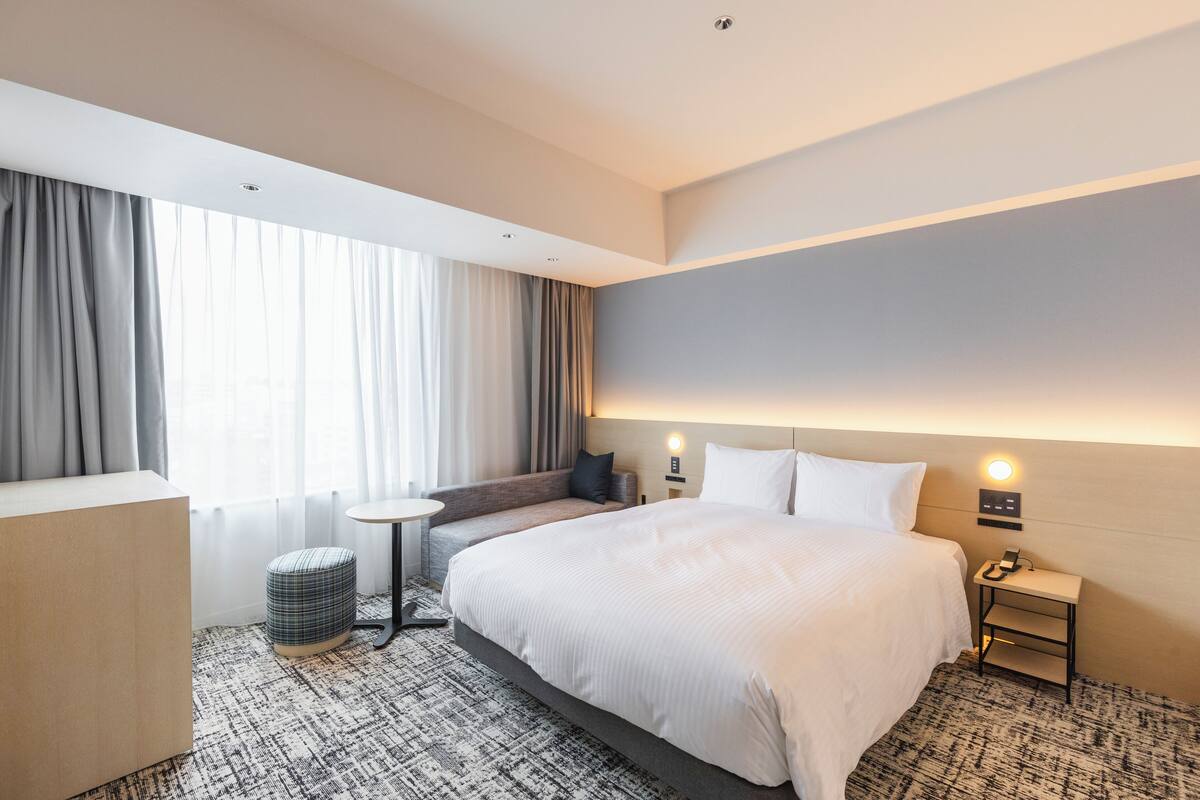 Una habitación de hotel con una cama grande, una mesita redonda pequeña, un sofá y una mesita de noche con una lámpara.