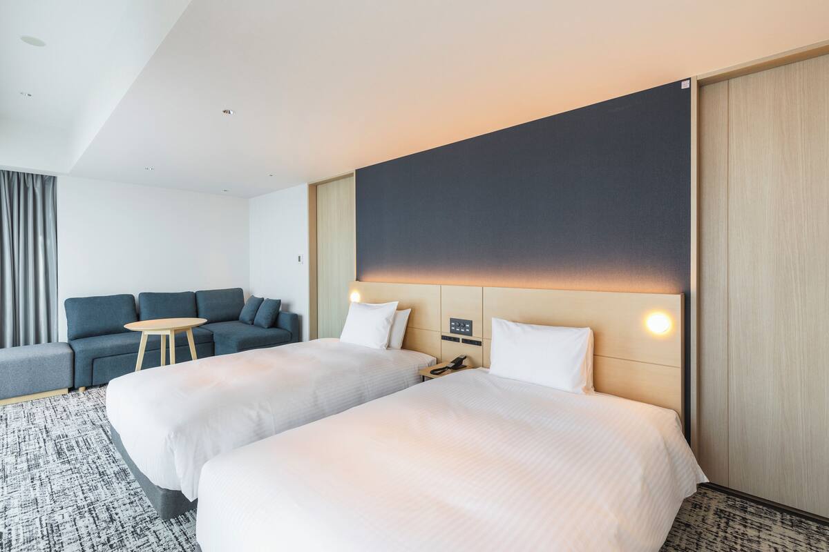 Habitación de hotel con dos camas, un sofá, una mesita y un televisor grande integrado en la pared.