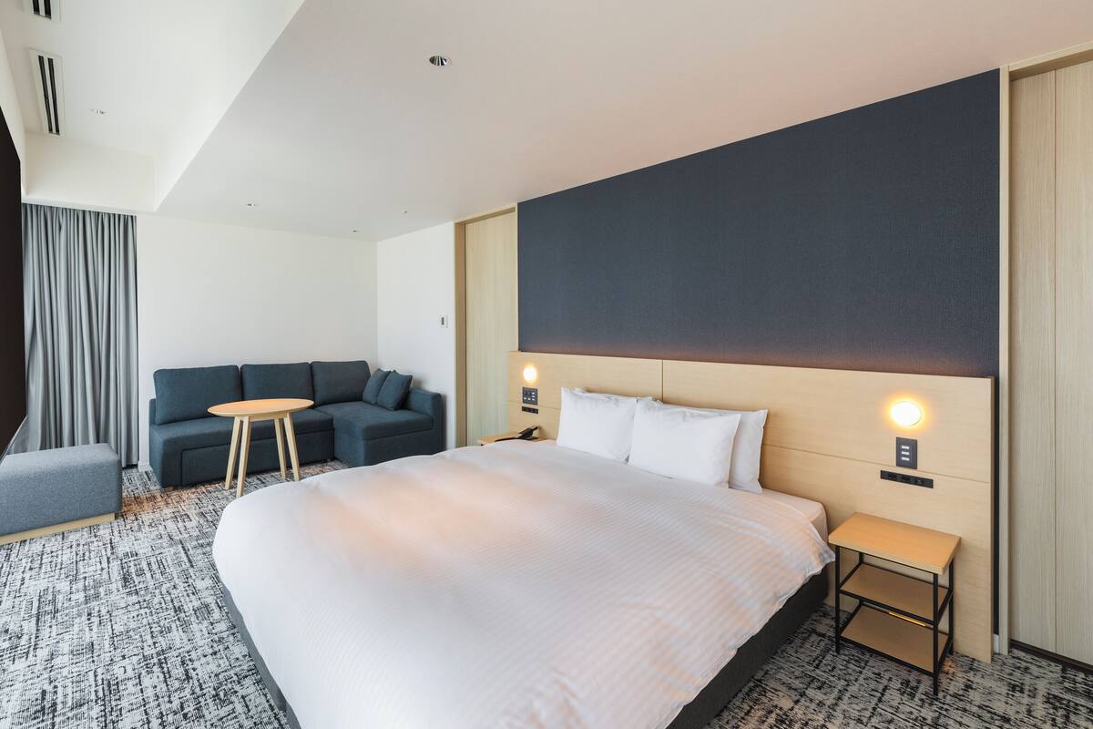 Una habitación de hotel moderna con una cama grande, un sofá, una mesita pequeña y una mesita de noche.