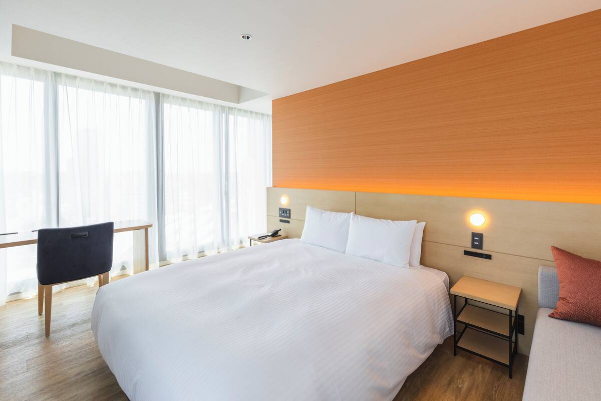 Habitación de hotel moderna con una cama grande, un escritorio y una silla. Se observa un mesita de noche con una lámpara y un teléfono. La habitación tiene un cabecero de madera y vistas a la ciudad a través de grandes ventanales.