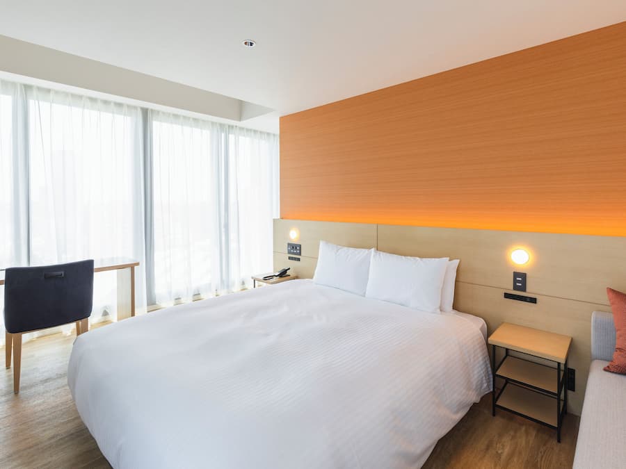 Une chambre d’hôtel moderne, dotée d’un grand lit, d’un bureau et d’une chaise. On y trouve une table de chevet avec une lampe et un téléphone. La chambre est agrémentée d’une tête de lit en bois et offre une vue sur la ville grâce à de grandes fenêtres.