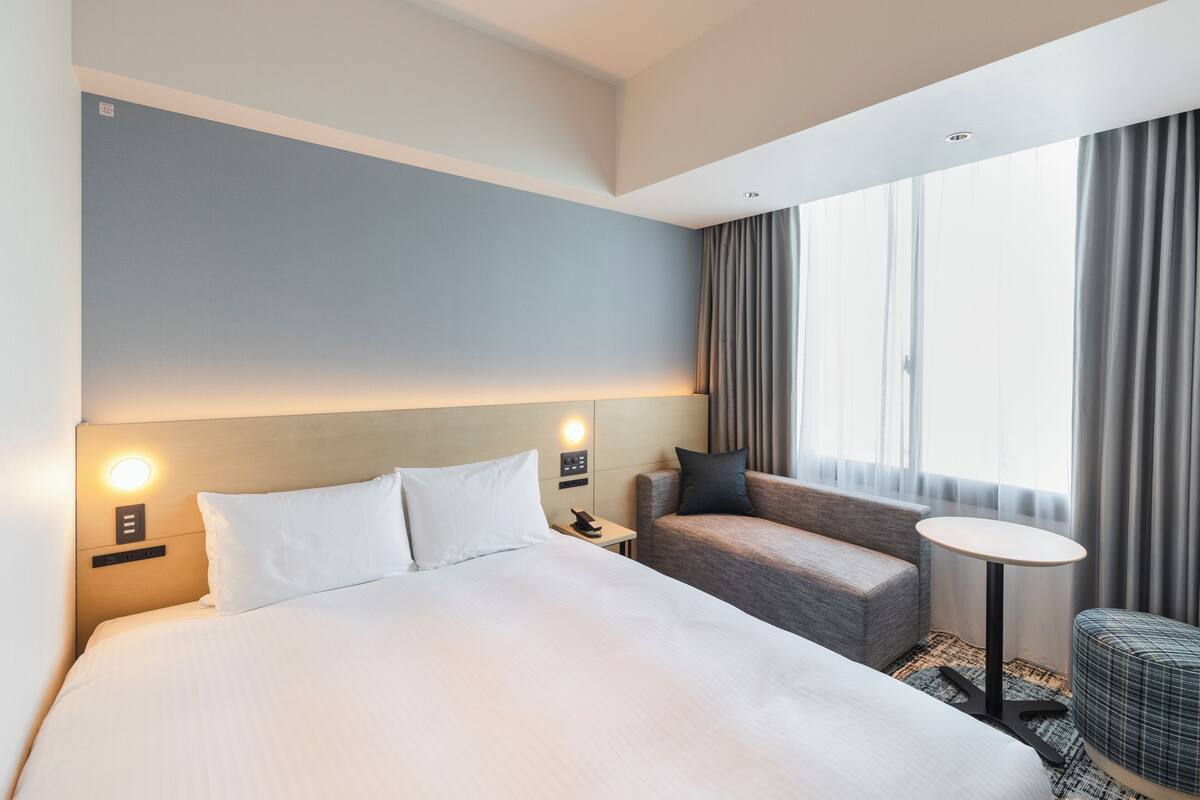 Una habitación de hotel con una cama grande, un sofá, una mesita y una ventana con cortinas.