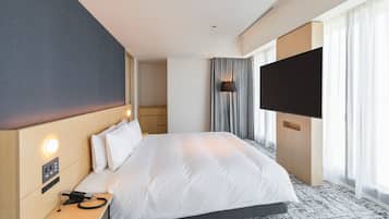 Ein Hotelzimmer mit einem großen Bett, einem Nachttisch mit Telefon und Ladegerät, einem flachbildfernseher an der Wand und einem Fenster mit Vorhängen.