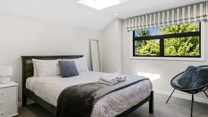A bedroom with a bed, a chair, a nightstand, a window with a view of trees, and a skylight. - Lake Taupō Hideaway Apartment in Acacia Bay (Lake Taupō)