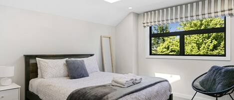 Un dormitorio con cama, una silla, una mesita de noche, un ventanal con vistas a árboles y un tragaluz.