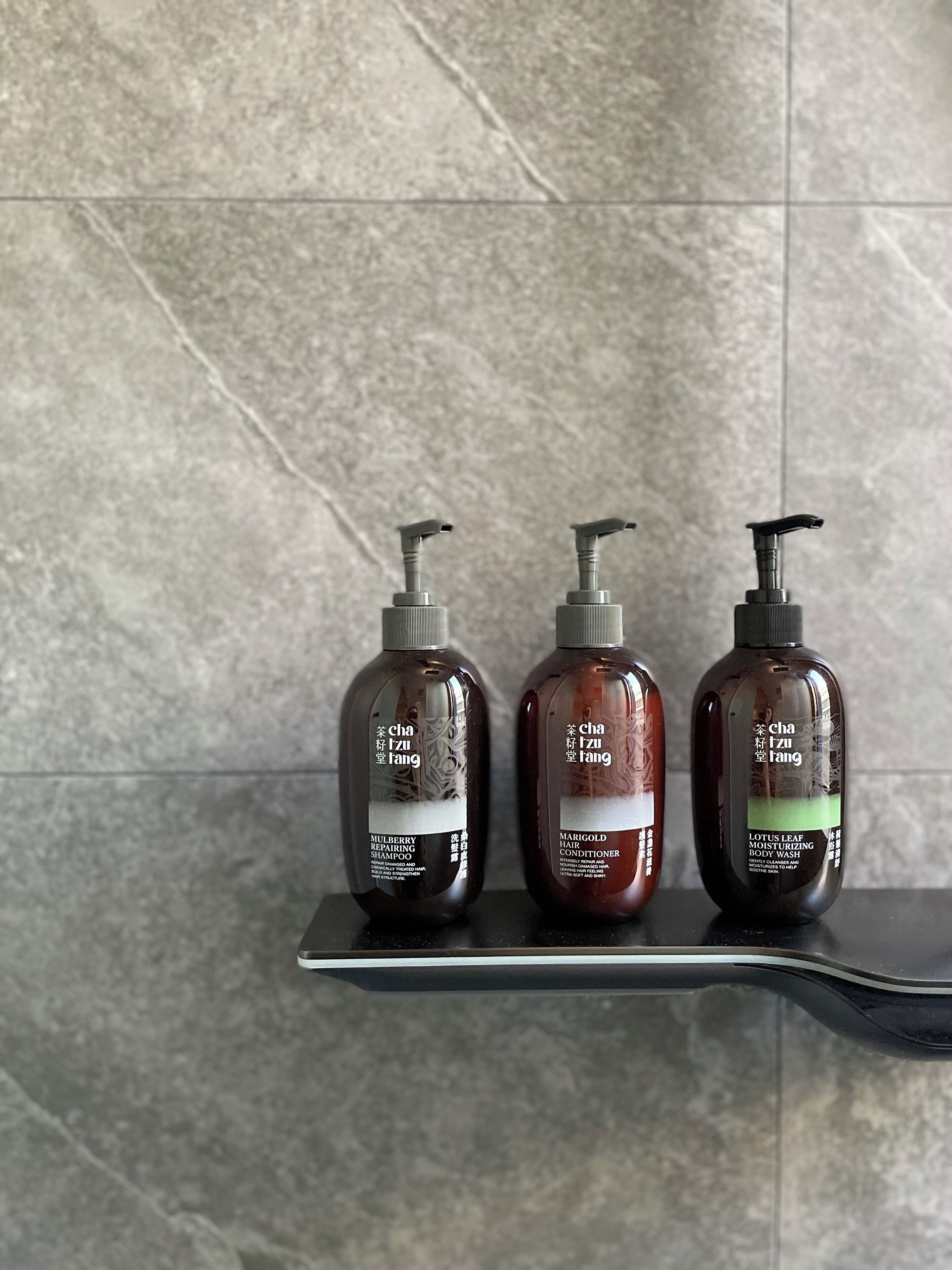 Tres botellas de productos capilares sobre una estantería frente a una pared con azulejos.