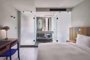 Ein modernes Hotelzimmer mit Schreibtisch aus Holz, blauem Sessel, Bett mit weißer Bettwäsche, Nachttisch aus Holz sowie einem Badezimmer mit Spiegel, Waschbecken und Handtuchhalter.