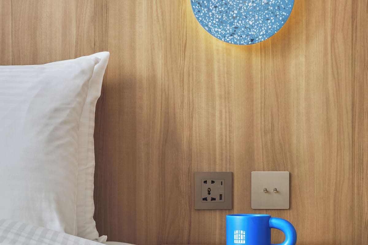 A neatly made bed with white linens, a wooden nightstand with a blue mug, and a wall socket.