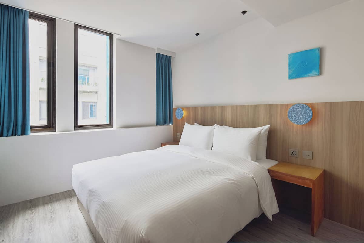 A hotel room with a large bed, wooden headboard, a nightstand, and a view of buildings through the window.