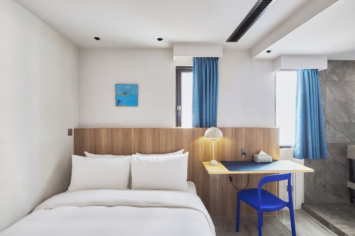 A modern hotel room with a bed, a desk, a chair, and a bathroom visible through an open door.