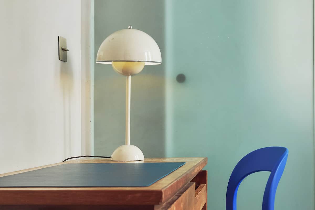 A wooden table with a white lamp and a blue chair in a room with a light green wall.