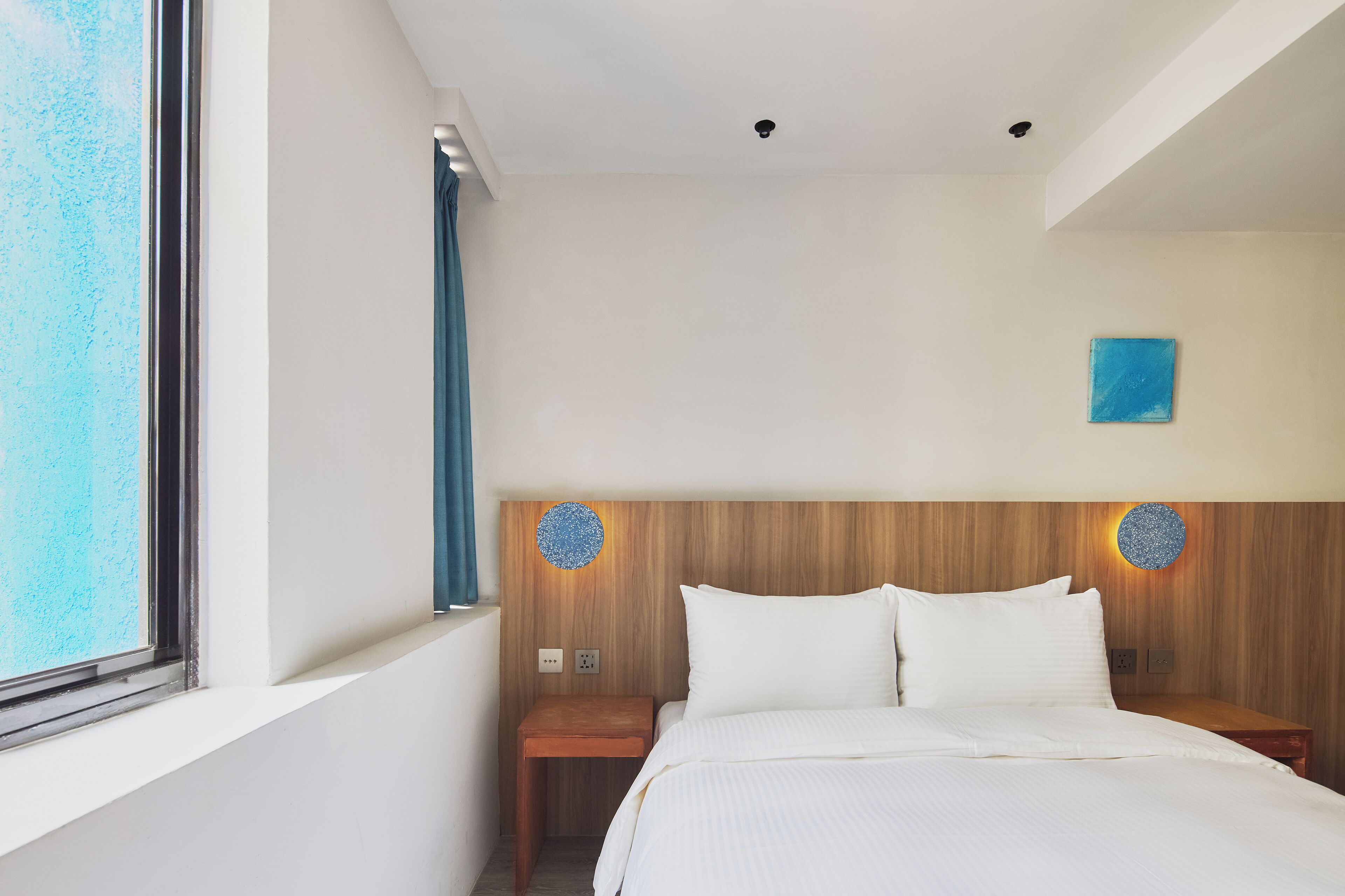 Habitación de hotel con una cama grande, cabecero de madera, dos lámparas de pared, una mesita de noche pequeña y una ventana con cortinas.