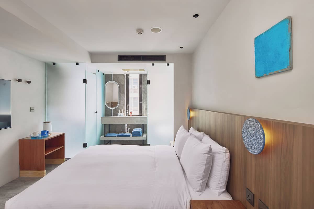 A modern hotel room with a large bed, a wooden headboard, a small wooden side table, a mirror, and a blue artwork on the wall.