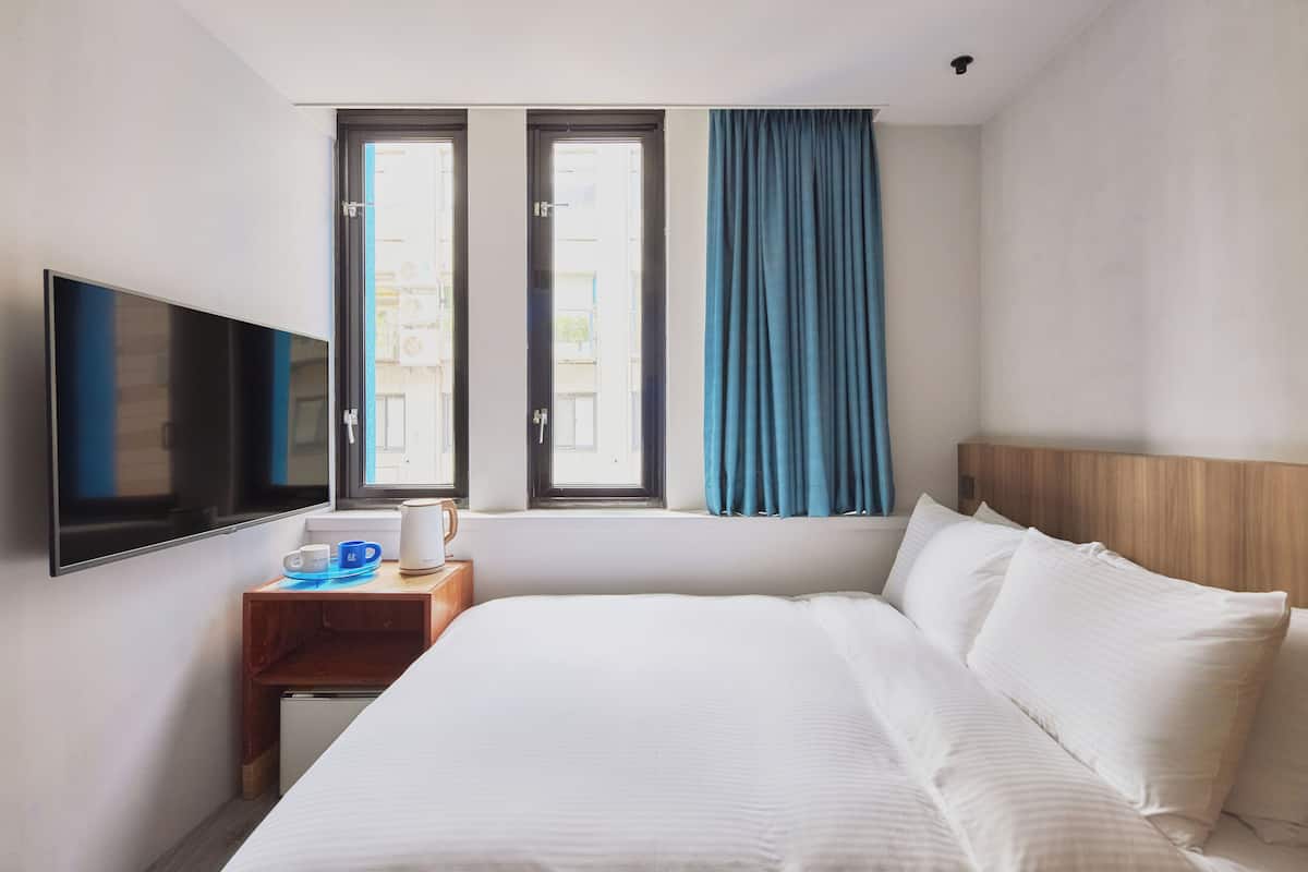 A hotel room with a bed, a TV, a window with curtains, and a small table with a cup and saucer.