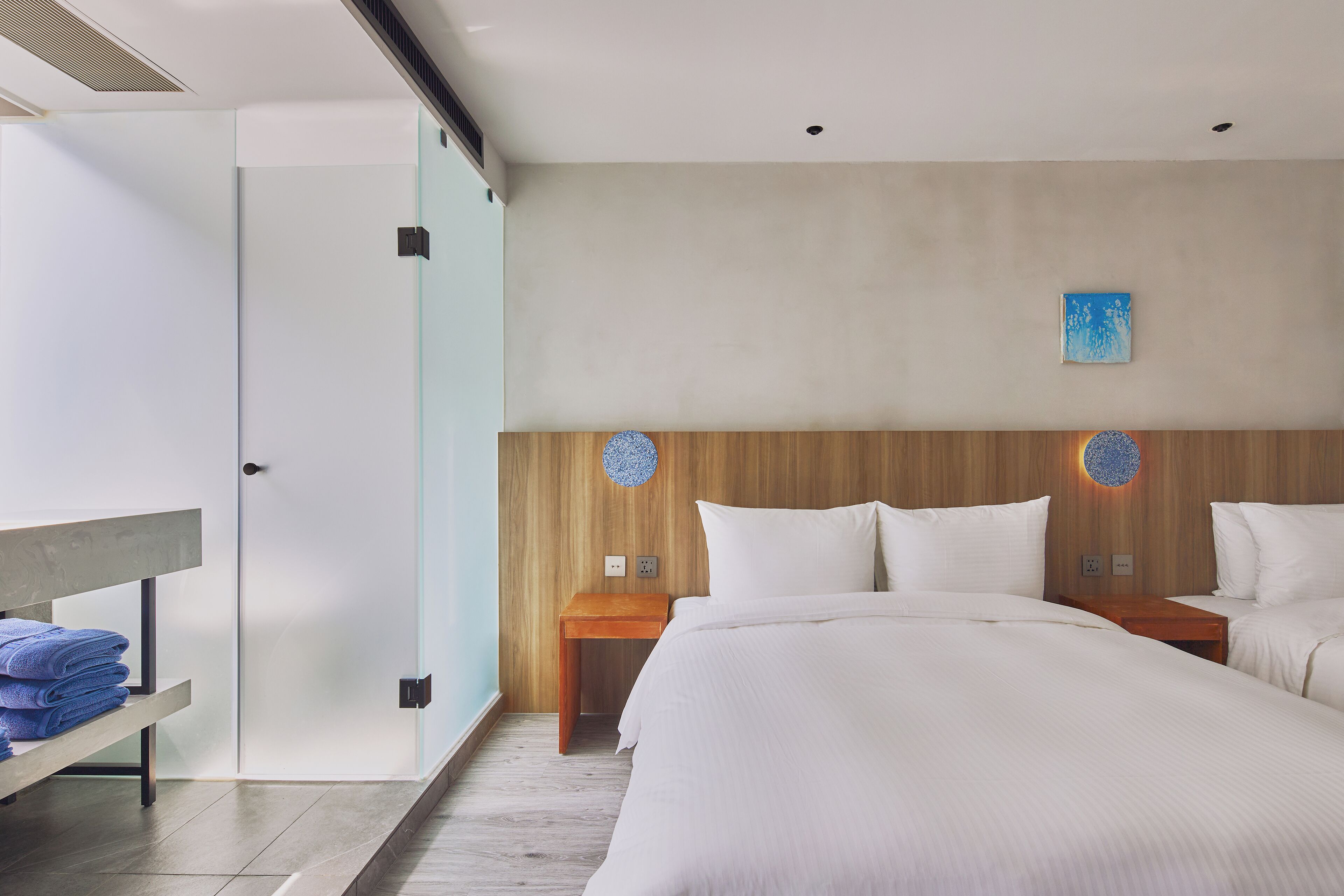 A modern hotel room with a large bed, a bedside table, a bench with towels, and a wooden headboard.