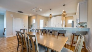 Un vaste espace de salle à manger avec une longue table en bois et des chaises assorties, une cuisine dotée de meubles blancs et d’un éclairage suspendu.