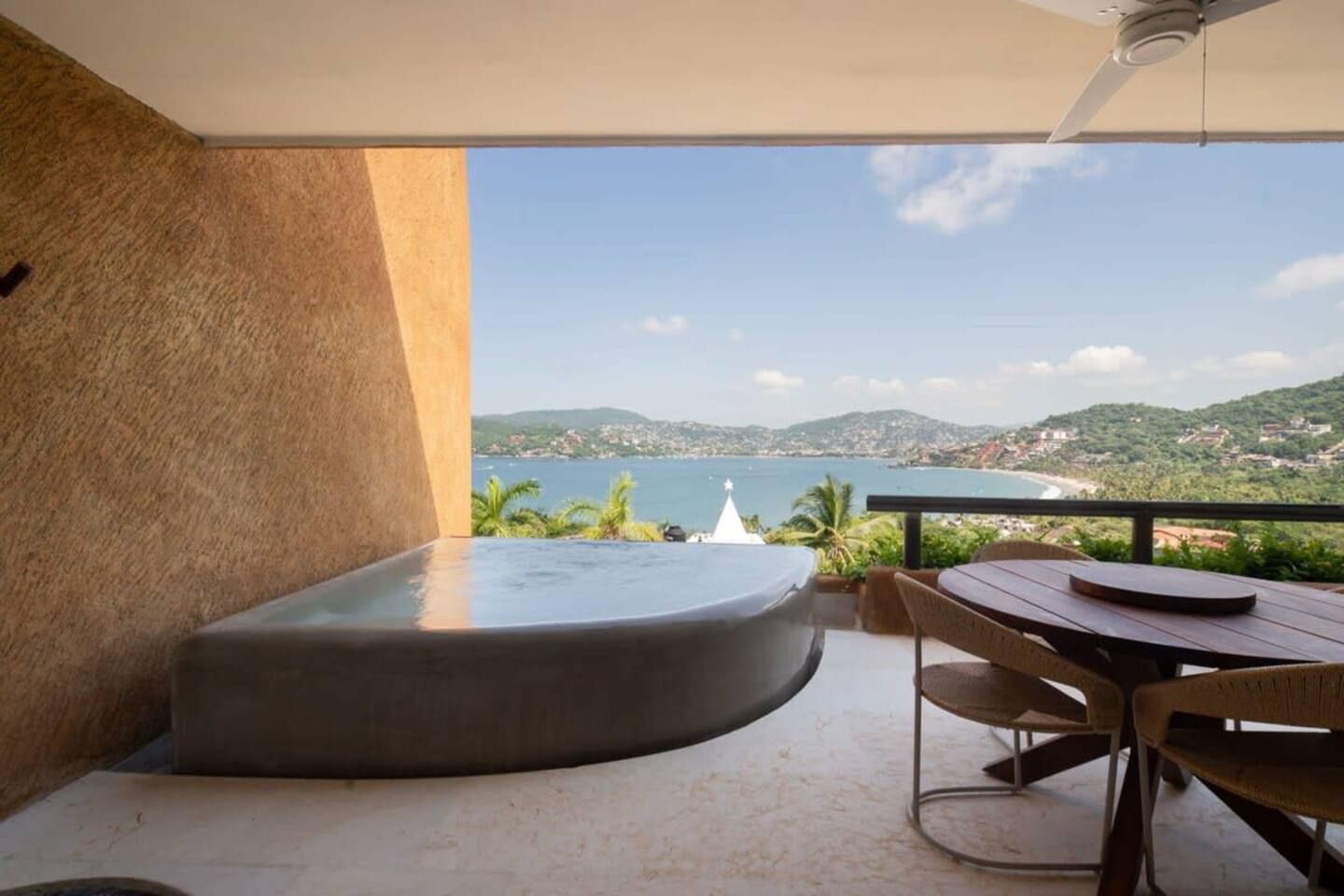 A balcony with a view of the sea and hills, a round seating area, and a table with chairs.