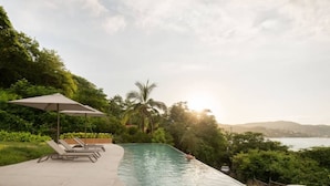 Piscina com espreguiçadeiras e um guarda-sol, cercada por vegetação exuberante e com vista para um corpo d'água.