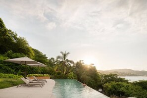 A swimming pool with lounge chairs and an umbrella, surrounded by lush greenery and overlooking a body of water. - Luxury Penthouse | Private Pool Steps from La Ropa (Zihuatanejo)