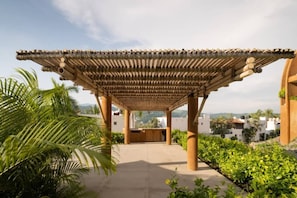 A covered outdoor area with a pergola structure, surrounded by greenery and overlooking a residential area.