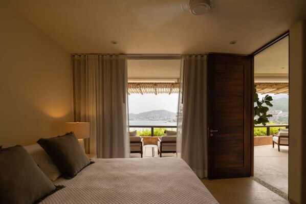 A bedroom with a large bed, a view of the outdoors, and a balcony with seating.