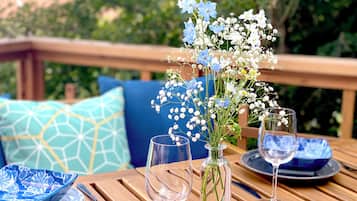 A wooden deck with a table set for dining, featuring a clear glass vase with flowers, two wine glasses, and a blue-patterned bowl.