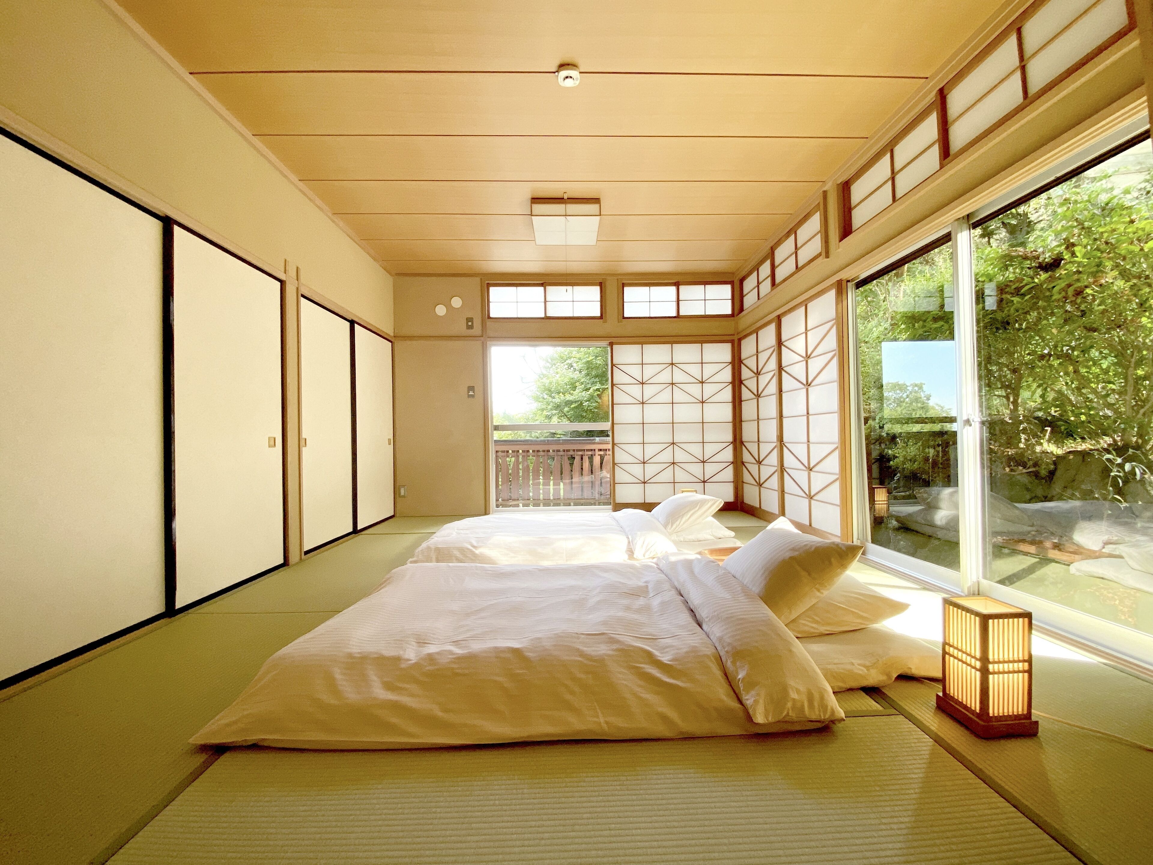 A traditional Japanese-style room with tatami flooring, a low bed with pillows, sliding shoji doors, and a view of greenery outside.
