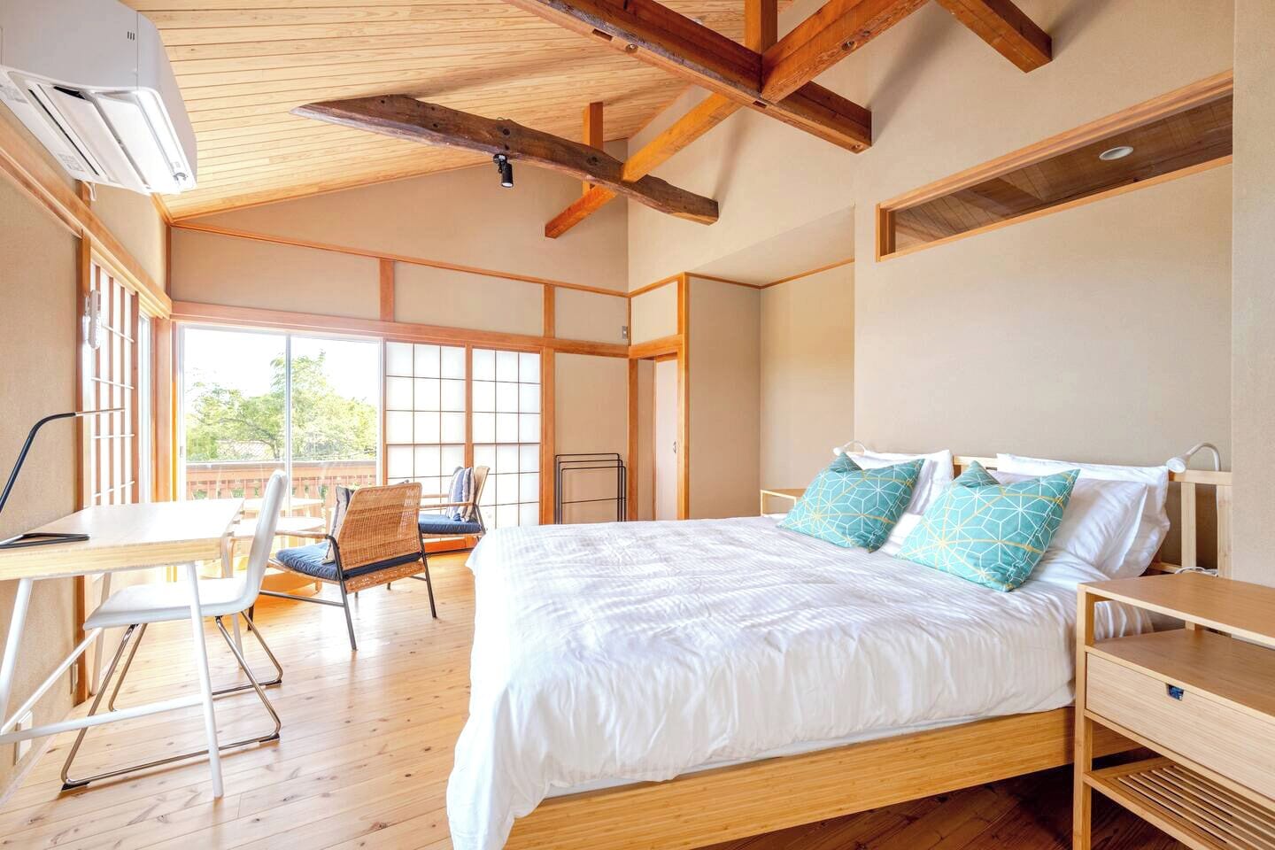 A bedroom with a wooden bed, a desk, and a chair. There is a window with a view of trees.