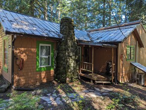 A rustic cabin with a stone chimney, a green window, and a porch with a wooden railing.