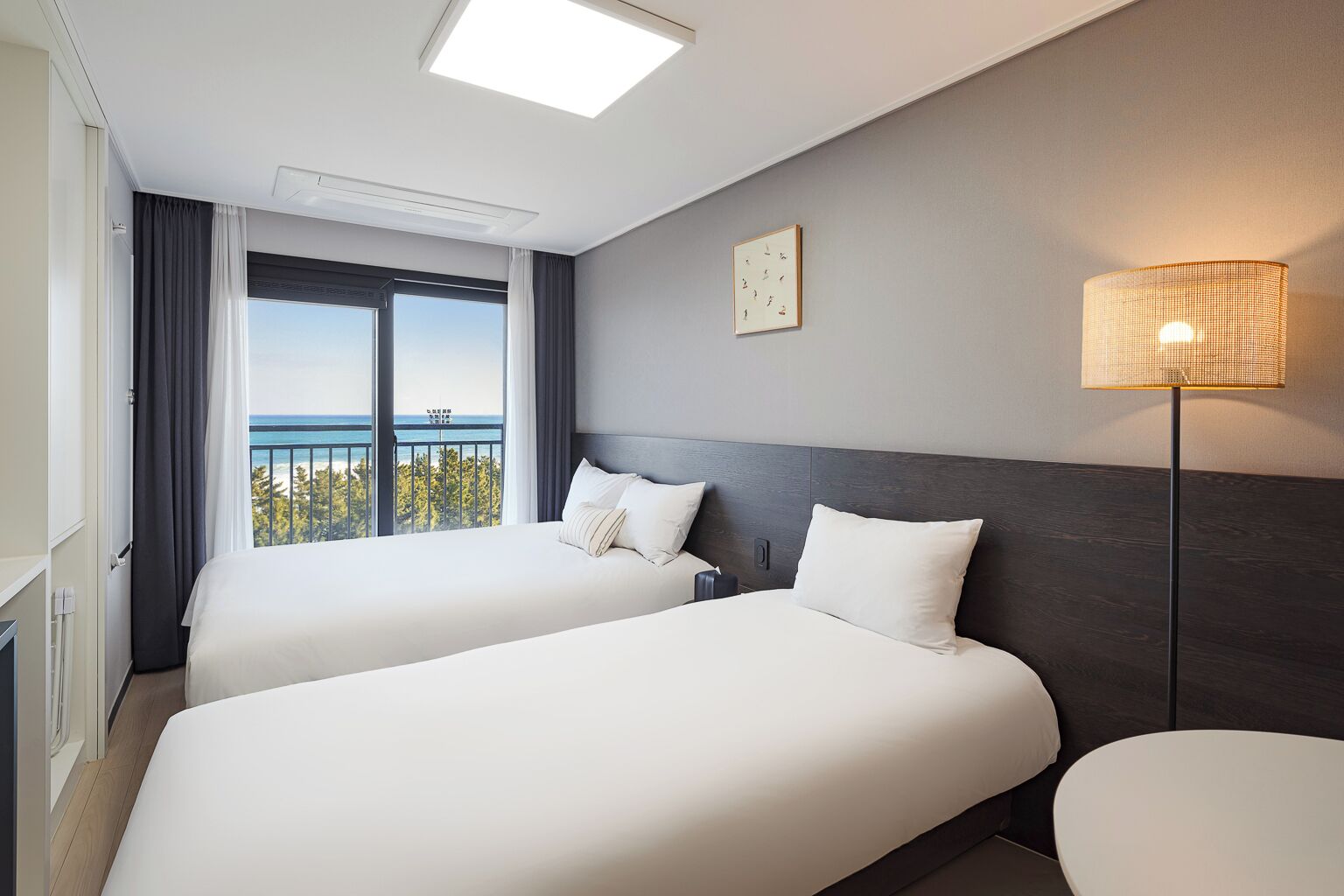 A hotel room with two beds, a view of the sea, a ceiling light, and a wall-mounted lamp.