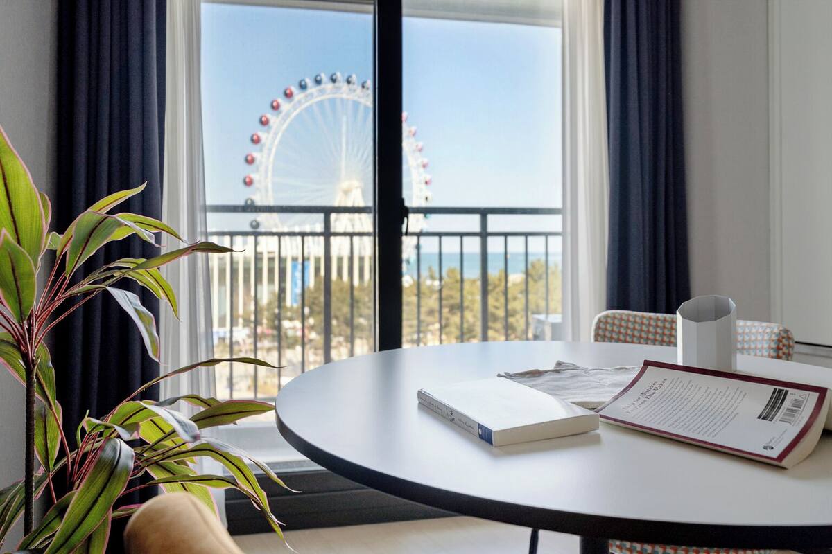 Ein Zimmer mit Blick auf ein Riesenrad, ein runder Tisch mit Buch und Speisekarte sowie eine Topfpflanze.