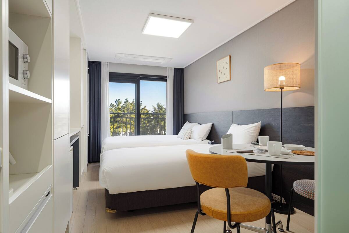 Ein Hotelzimmer mit einem Bett, einem Schreibtisch mit Lampe, einem Sessel und Blick auf Bäume durch das Fenster.