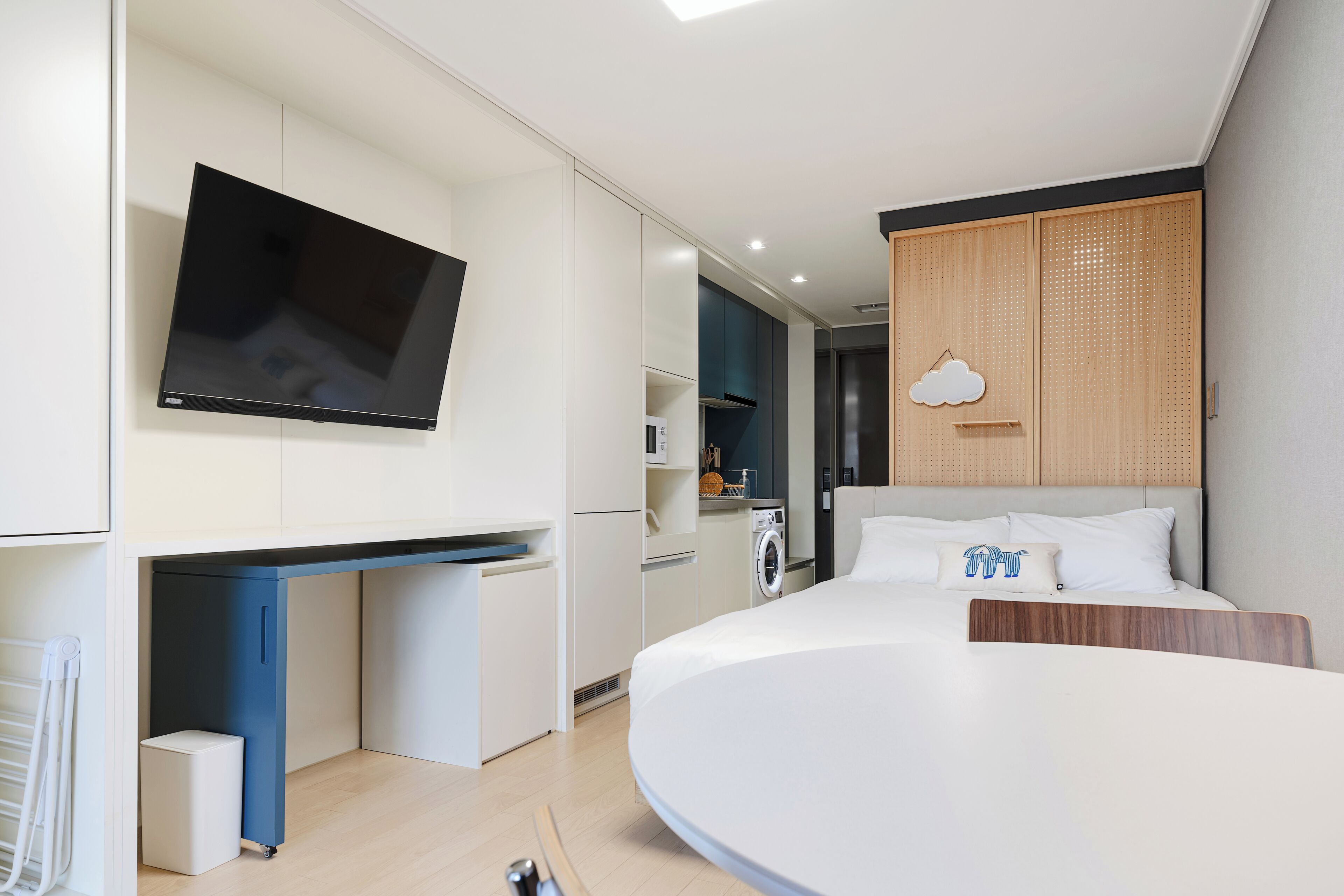 A modern hotel room with a flat-screen TV mounted on the wall, a compact kitchenette, a bed with a wooden headboard, and a ceiling-mounted light fixture.